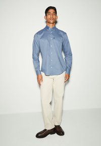 Camicia azzurra con bottoni, con un piccolo motivo geometrico, maniche lunghe e colletto; abbinata a pantaloni crema e scarpe marroni.