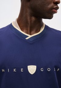 Άτομο με σκούρο δέρμα φορεί μια μπλε σκουρόχρωμη μπλούζα Nike Golf με μπεζ γιακά και κεντημένο λογότυπο στο στήθος.