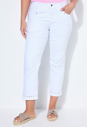 Kobieta w białych skróconych dżinsach z ozdobnymi przelotkami na końcach i beżowych sandałach wsuwanych, stoi z jedną ręką w kieszeni.