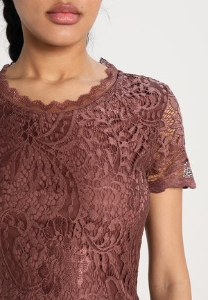 Femme portant un haut à manches courtes en dentelle mauve avec des motifs floraux et un col festonné sur un fond uni.