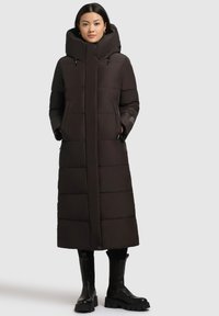 Manteau long rembourré à capuche de couleur marron foncé avec un motif matelassé, doté de deux poches avant et d'une coupe droite, associé à des bottes noires hauteur genou.