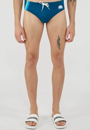 Homme portant un slip de bain bleu avec des rayures vertes et bleu clair, un cordon blanc, des claquettes blanches et des tatouages visibles sur les jambes sur fond uni.