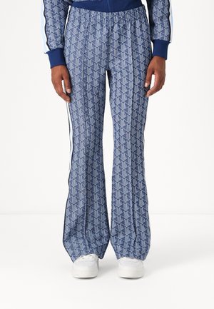 Siniset kuviolliset liehuhousut valkoisilla sivuraitoja, valkoisten tennareiden kanssa, kädet rentona sivuilla valkoista taustaa vasten.