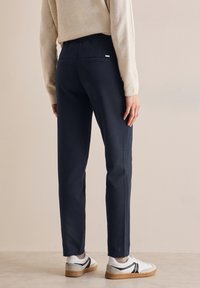 Navy trousers featuring a fitted design, smooth texture, and back pockets. Worn with white sneakers and a beige sweater.