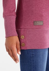 Mano appoggiata accanto a una maglietta rosa a maniche lunghe con due bottoni decorativi e un piccolo logo, abbinata a jeans in denim blu.