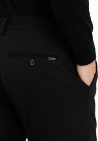 Schwarze Hosen mit einer Gesäßtasche, die mit einem Knopfverschluss und einem kleinen metallischen Akzent ausgestattet ist. Der Stoff hat eine glatte, leicht strukturierte Optik.