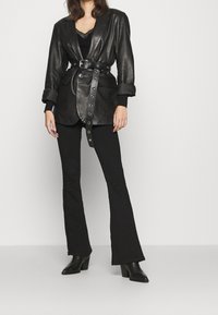 Blazer en cuir noir à coupe cintrée, taille marquée et accessoires argentés. Associé à un pantalon évasé noir et des bottines à talons.