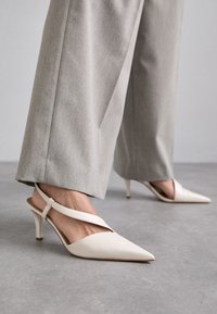 White leather slingback heels with a pointed toe, slim heel, and crisscross strap design. Worn with light gray wide-leg trousers.