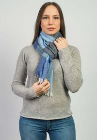 Mnohobarevný šátek v odstínech modré s texturami a vzory. Dokončený třásněmi. Nosený přes šedý svetr a modré džíny.