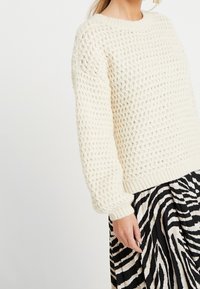 Dicker gestrickter cremfarbener Pullover mit einem strukturierten Gewebe-Muster, rundem Ausschnitt und gerippten Bündchen, kombiniert mit einem kontrastierenden schwarz-weißen gestreiften Rock.