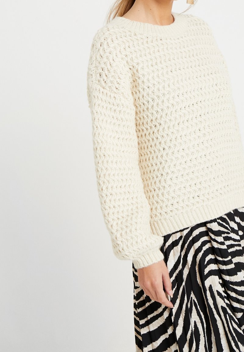Dicker gestrickter cremfarbener Pullover mit einem strukturierten Gewebe-Muster, rundem Ausschnitt und gerippten Bündchen, kombiniert mit einem kontrastierenden schwarz-weißen gestreiften Rock.