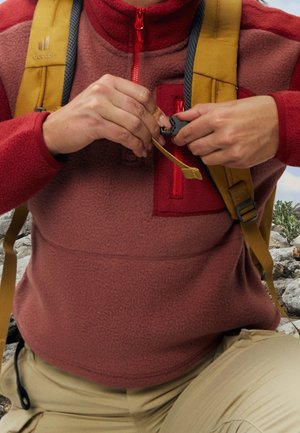 Personne en veste polaire rouge attachant la sangle jaune d'un sac à dos en extérieur sur un terrain rocheux.