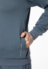 Blauwe sweatshirt met een zachte textuur, voorzien van een zijzak en de tekst "SHORT STORIES" in het wit op de heup. Ronde halslijn en geribbelde manchetten.