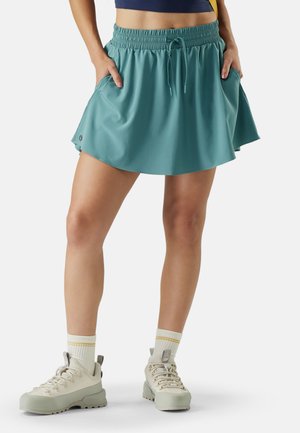 Femme portant une skort de sport couleur sarcelle avec taille à cordon et poches avant, associée à des chaussures de randonnée beige et des chaussettes blanches à rayures jaunes.