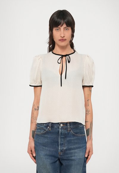 Blouse à manches courtes couleur crème clair à pois noirs, encolure goutte d'eau et bordure noire. Caractérisée par des manches bouffantes et un tissu léger.