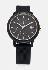 Reloj con correa de silicona negra, esfera redonda negra, marcadores de horas dorados, tres subesferas y un logotipo dorado de cocodrilo.