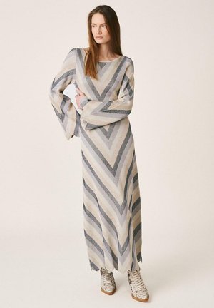 Mujer con un vestido largo de punto con patrón de chevron a rayas y mangas anchas, combinando con botines de piel de serpiente, de pie con un brazo cruzado.