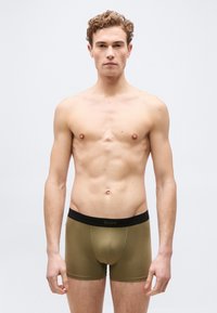 Νέος άνδρας με κοντά, σγουρά μαλλιά, φορώντας εσώρουχα boxer σε ελιά πράσινο με μαύρη ζώνη μέσης, στέκεται μπροστά σε απλό φωτεινό φόντο.