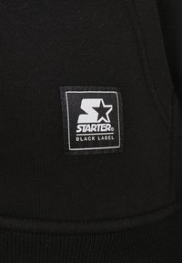Sort hættejakke med struktureret stof, prydet med et firkantet hvidt mærke. Mærket inkluderer "STARTER" og et stjerne-logo, broderet på fronten.