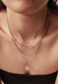 Drielaagse ketting in rosegoud met ronde en ovale kralen, met een ronde hanger die hangt aan de laagste ketting.