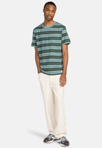 Striped T-shirt in teal and dark green with short sleeves and a chest pocket, paired with loose off-white pants and multi-color sneakers.
