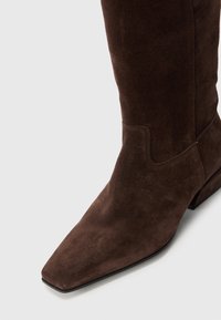 Botte en suède marron, montant jusqu'au genou, avec un bout carré et un petit talon, présentant une texture lisse et des détails de couture minimaux.