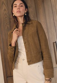 Giacca cropped in suede verde oliva con grandi bottoni, tasca sul petto e una texture liscia, abbinata a un top bianco a righe e pantaloni beige.