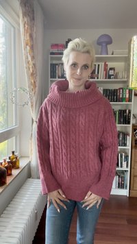 Dikke kabelgebreide trui in rozenroze, met een brede kraag en lange mouwen. Gecombineerd met blauwe jeans, gesitueerd in een gezellige interieur.