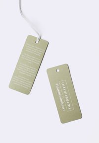 Groene papieren labels met witte tekst; de voorkant toont de merknaam "#intimissimicares" en de achterkant bevat meertalige details over de samenstelling van de stof.