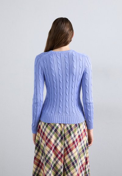 Light blue cable-knit sweater with long sleeves and ribbed hem, worn over a plaid skirt featuring various colors including green, red, and yellow.
