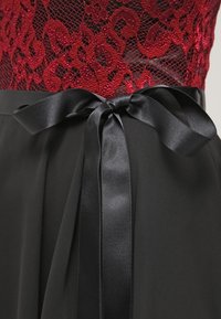 Robe noire avec un corsage en dentelle rouge orné de motifs floraux et un ruban de taille noir en satin noué en nœud, mettant en avant son design élégant.
