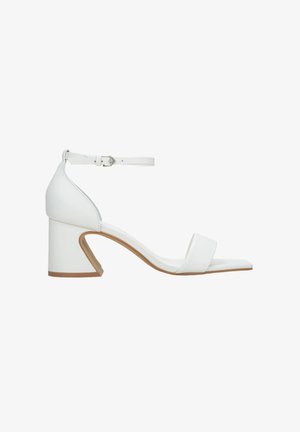 White leather heeled sandals with an ankle strap, square toe, and a unique triangular block heel design. Smooth texture and minimalist style.