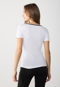 Balta trumpa rankovė t-shirt, banguota tekstūra, tamsiai mėlyna apykaklė, prigludęs dizainas. Fonas paprastas, pabrėžiantis marškinėlių ypatybes.