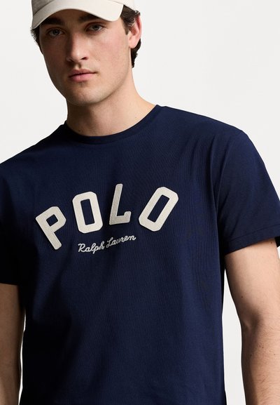 Camiseta de algodón azul marino con un gran gráfico en crema que dice "POLO" y el texto "Ralph Lauren" debajo, cuello redondo y mangas cortas.