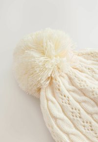 Gehaakte crèmekleurige beanie met een grote, fluffy pompon bovenop, met een getextureerd kabelpatroon over het hele hoofdgedeelte.