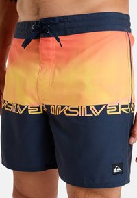 Herrbadshorts med ett orange till gult gradiente mönster upptill, med en marinblå nederdel och Quiksilver-logotyp i gult.