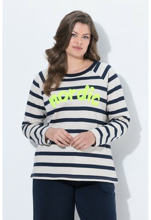 Donna con lunghi capelli castani indossa una maglietta a maniche lunghe bianca e a righe blu navy con "nordic" in giallo e pantaloni blu scuro.