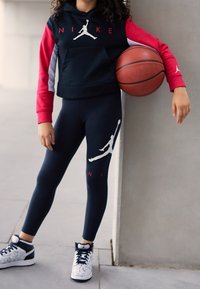 Bambino in abbigliamento Nike nero e rosso con una palla da basket, appoggiato a un pilone in un ambiente urbano all'aperto.