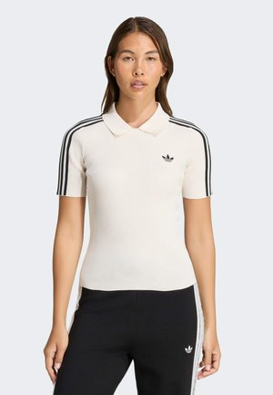 Donna che indossa una polo bianca a maniche corte con strisce nere sulle maniche e pantaloni neri con una striscia bianca sul lato, entrambi del marchio Adidas.