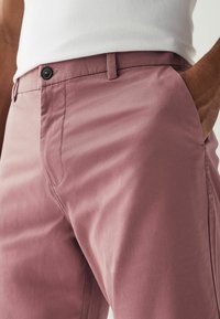 Lyserøde chinos lavet af blødt stof, med en enkelt knaplukning, sidelommer og et strømlinet design med synlige sømme.