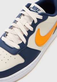 Baskets Nike avec une tige en cuir beige, accents marine, logo orange et lacets blancs. Comprend un bout arrondi et des détails perforés.