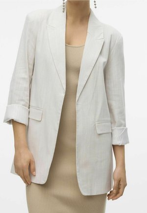 Kvinde iført en lys beige ribbet kjole og en hvid blazer med opkrullede ærmer mod en ensfarvet hvid baggrund.