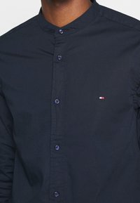 Camicia blu con colletto alla mandarino, con bottoni scuri e un piccolo logo rosso, bianco e blu sul lato sinistro del petto. Tessuto di texture liscia.