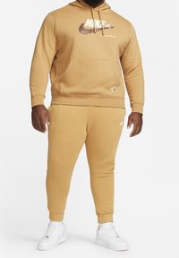 Nike mikina a tepláky v teplé béžové barvě, vyrobené z měkké látky. Mikina má grafické logo a přední kapsu. Bílé tenisky.