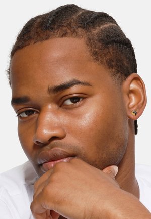 Close-up of a young man with braided hair, sporting a gold stud earring. He has smooth skin and a neutral shirt, resting his chin on his hand.