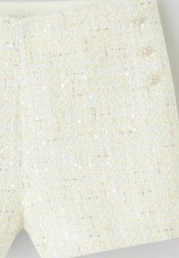 Shorts en tweed crème avec un tissu texturé orné de sequins et trois boutons décoratifs ressemblant à des perles sur la ceinture.