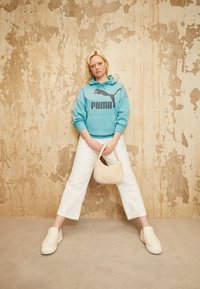 Blondýnka stojí s rozkročenýma nohama, na sobě má modrou mikinu Puma, bílé kalhoty, v ruce drží béžovou kabelku a má na sobě bílé slip-on boty, před texturovanou zdí.