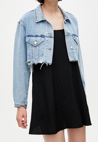 Vaaleansininen kulutettu denim-takki, jossa on kaksi rintataskua, napitus ja rispaantunut helma. Päällä musta ilmanhihoja mekko.