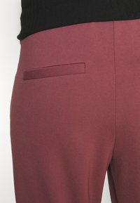 Burgundijske hlače s glatkom, teksturiranom završnom obradom, koje imaju jednu stražnju džep s ravnim dizajnom. Pojas je crn i rebrast.