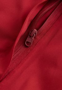 Röd tyg med en stängd matchande röd dragkedja och dragflik i närbild, vilket visar detaljerad sömnad och textur.